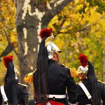 Des soldats, coiffés de casques ornés de longues plumes noires, se tiennent en formation au milieu des arbres d'automne. Leurs uniformes et leurs touches de rouge confèrent un caractère formel et cérémoniel.