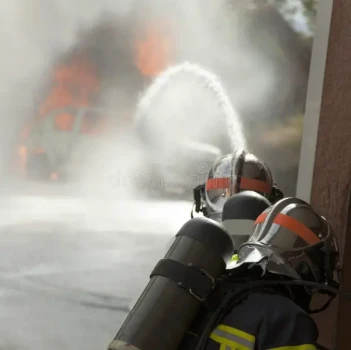 Des pompiers, casqués et équipés de protection, pointent une lance d'incendie sur un véhicule enfumé et en flammes. La scène illustre l'urgence et le travail d'équipe des pompiers.