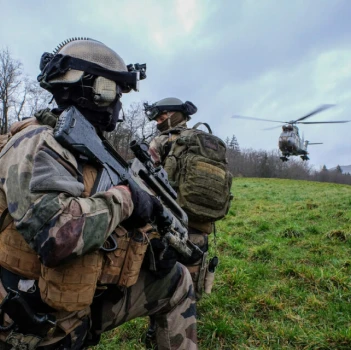 Deux soldats en tenue de camouflage et casques, équipés de fusils, s'agenouillent sur un terrain herbeux alors qu'un hélicoptère s'approche, transmettant une scène tendue et pleine d'action.