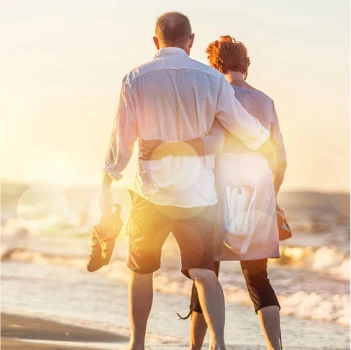 Un couple marche bras dessus bras dessous sur une plage ensoleillée, éclairée par le coucher de soleil doré. Ils portent des chaussures, évoquant chaleur, détente et complicité.