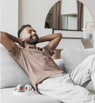 Un homme se détend sur un canapé gris, souriant, les mains derrière la tête. Un casque audio est posé à côté de lui. La pièce est moderne, calme et baignée de lumière tamisée.