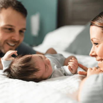 Un couple souriant est allongé sur un lit, contemplant avec amour leur nouveau-né. La lumière tamisée et les expressions chaleureuses créent une atmosphère intime et joyeuse.