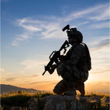 Silhouette d'un soldat agenouillé avec un fusil au coucher du soleil, au sommet d'une colline herbeuse, ciel aux teintes orange et bleues. La scène respire la vigilance et le calme.