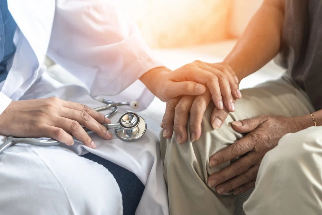 Un professionnel de santé tient délicatement la main d'un patient âgé pour le réconforter. Un stéthoscope est posé à proximité. La scène exprime bienveillance et empathie.