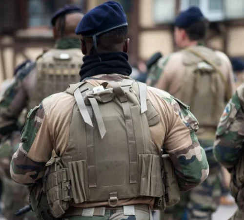 A group of soldiers in camo uniforms and berets walk together, backs facing the camera. They carry heavy backpacks, conveying a sense of readiness.