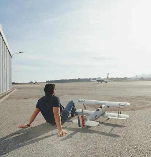 Une personne est assise sur une piste d'atterrissage, adossée à un biplan miniature. Au loin, un gros jet se prépare au décollage. La scène est sereine et contemplative.
