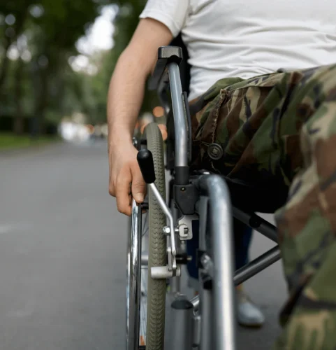 Une personne en pantalon camouflage et chemise blanche se déplace en fauteuil roulant sur un sentier bordé d'arbres. L'image évoque le mouvement et l'indépendance.