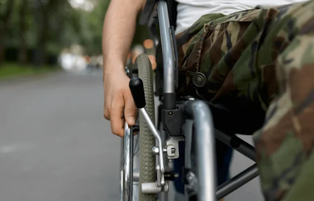 Une personne en pantalon camouflage et chemise blanche se déplace en fauteuil roulant sur un sentier bordé d'arbres. L'image évoque le mouvement et l'indépendance.