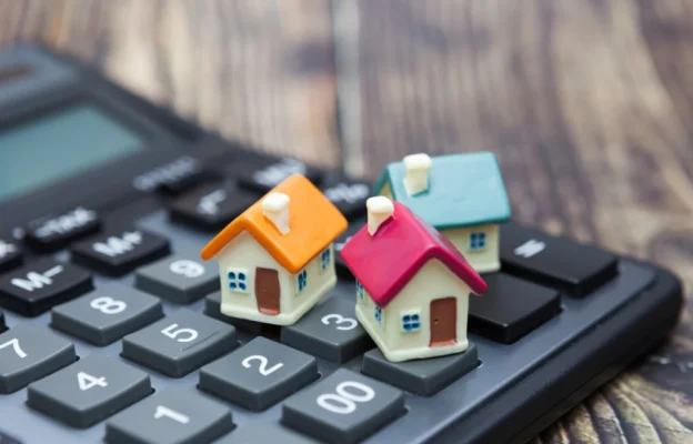Trois maisons colorées posées sur une calculatrice symbolisent les coûts de l'immobilier. Le fond en bois met en valeur le thème de la planification financière.