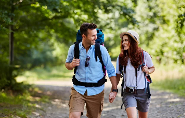 Un couple souriant marche sur un sentier forestier, main dans la main. Ils portent des sacs à dos et des vêtements d'été décontractés, exprimant un sentiment d'aventure et de bonheur.