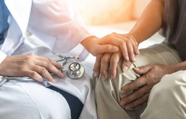 Un professionnel de santé tient délicatement la main d'un patient âgé pour le réconforter. Un stéthoscope est posé à proximité. La scène exprime bienveillance et empathie.