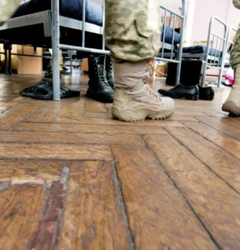 Des soldats en uniformes de camouflage et bottes beiges marchent sur un parquet ciré. Des lits et des chaises sont visibles, suggérant une ambiance de caserne militaire.