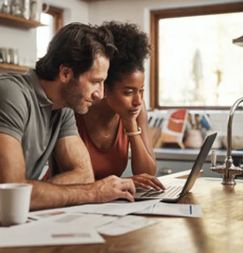 Un couple regarde attentivement un ordinateur portable dans une cuisine. Des papiers et une tasse à café sont posés sur la table en bois. L'ambiance est chaleureuse et conviviale.