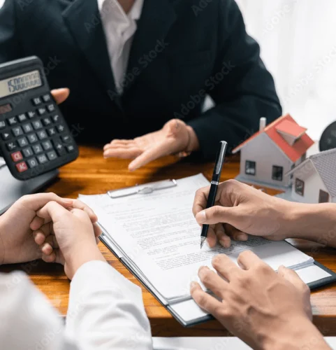 Une réunion d'affaires réunit trois personnes discutant d'un document. L'une tient un stylo, l'autre une calculatrice. Une maquette de petite maison symbolise l'immobilier.