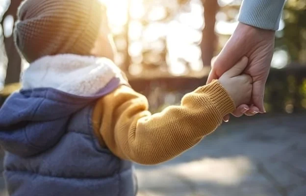 Un enfant vêtu d'une veste douillette et d'un bonnet tient la main d'un adulte. L'arrière-plan est légèrement flouté par la lumière vive du soleil, évoquant chaleur et sécurité.