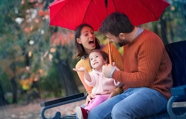 Une famille joyeuse est assise sur un banc sous un parapluie rouge, sous la pluie. L'enfant, vêtu d'une veste rose, lève les yeux avec émerveillement, tandis que les parents sourient et rient.
