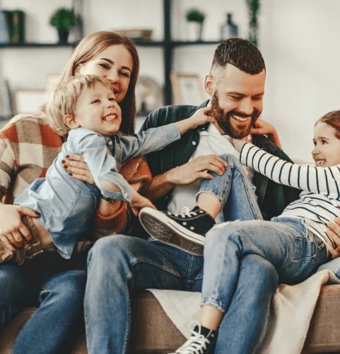 Une joyeuse famille de quatre personnes est assise sur un canapé, riant et jouant ensemble. Les parents serrent leurs jeunes fils et fille contre eux, créant une atmosphère chaleureuse et affectueuse.