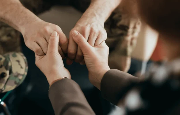 Une personne en uniforme militaire est assise, les mains tenues par une autre personne de manière encourageante, transmettant réconfort et empathie dans un cadre de conseil.