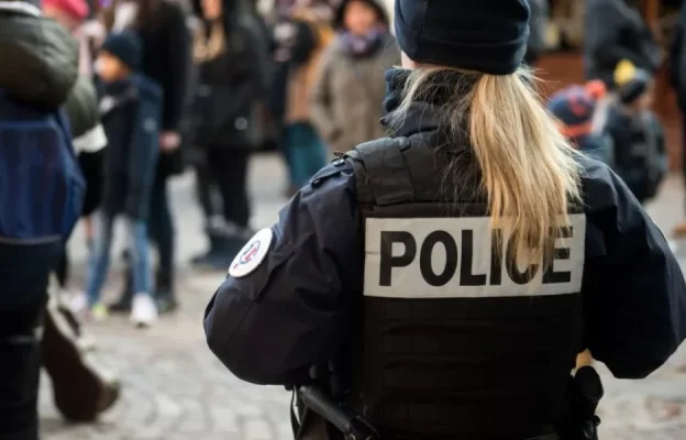 Un policier coiffé d'une queue de cheval se tient au milieu de la foule, vêtu d'une veste marquée « POLICE ». La scène suggère la présence des forces de l'ordre dans un espace public.