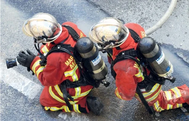 Deux pompiers en uniformes rouges et casques sont agenouillés sur l'asphalte mouillé, une lance à la main. Ils portent des appareils respiratoires, créant un sentiment d'urgence et de travail d'équipe.