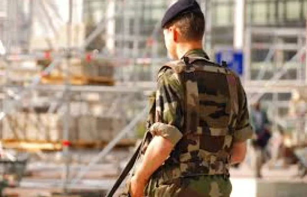 Un soldat en uniforme camouflage et béret se tient debout, un fusil sur le dos, surplombant un chantier de construction avec des échafaudages. La scène évoque la vigilance.