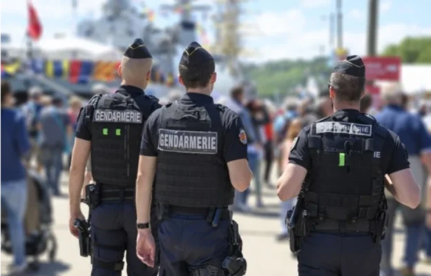 Trois gendarmes en uniforme traversent une manifestation en plein air bondée. Ils se détournent, surveillant la foule. Police et ambiance publique.