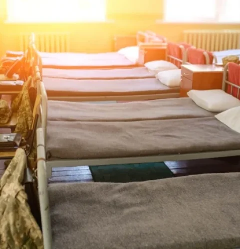 Une pièce ensoleillée avec plusieurs lits identiques, de style militaire, soigneusement faits et alignés. Chacun est doté d'une couverture grise, d'un oreiller blanc et d'un équipement de camouflage sur le côté, évoquant un sentiment de discipline.