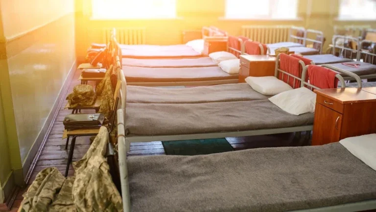 Une pièce ensoleillée avec plusieurs lits identiques, de style militaire, soigneusement faits et alignés. Chacun est doté d'une couverture grise, d'un oreiller blanc et d'un équipement de camouflage sur le côté, évoquant un sentiment de discipline.