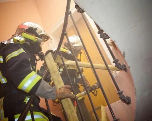 Deux pompiers en tenue complète, casque et appareil respiratoire inclus, montent un escalier enfumé, une lance à la main. L'atmosphère est tendue et l'urgence est palpable.