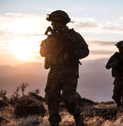 Des soldats en silhouette sur fond de soleil couchant marchent dans un paysage rocheux, évoquant un sentiment de détermination et de camaraderie.