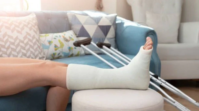 Une personne est allongée sur un canapé bleu, une jambe dans un plâtre blanc posée sur un coussin rond. Des béquilles sont à proximité et des oreillers créent une atmosphère chaleureuse.