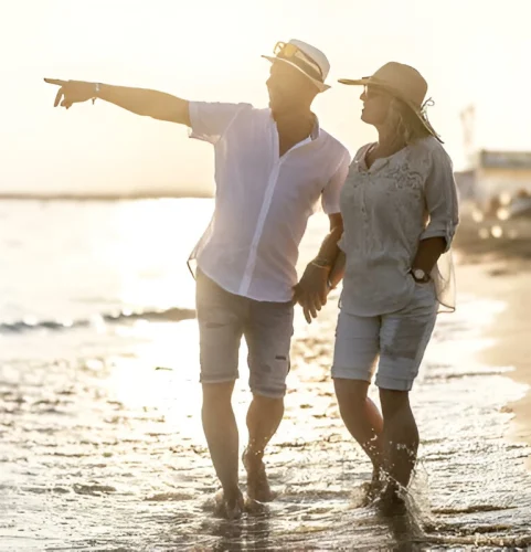 Un couple se promène sur une plage au coucher du soleil, main dans la main et sourire aux lèvres. L'homme pointe du doigt le lointain. La lumière chaude crée une ambiance sereine et joyeuse.