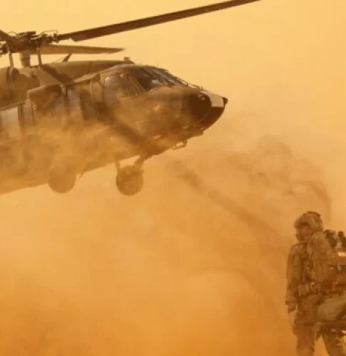 Un hélicoptère militaire plane près du sol, soulevant un nuage de poussière. Un soldat se tient à proximité, évoquant une atmosphère intense et trépidante.