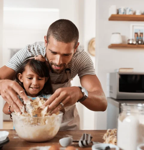 Un père et sa fille cuisinent joyeusement dans une cuisine. Ils mélangent la pâte dans un saladier en verre, entourés d'œufs et de farine, avec des étagères et un micro-ondes en arrière-plan.