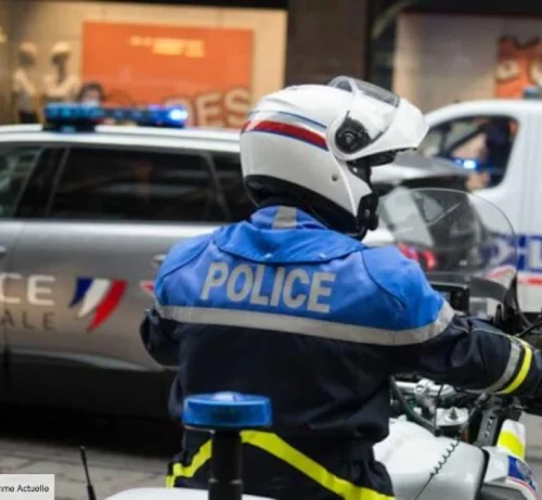 Un policier à moto, portant un uniforme bleu et un casque, est vu de dos. Une voiture et une camionnette de police équipées de gyrophares apparaissent à l'arrière-plan.